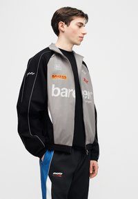 Jeune homme portant une veste sportive noire, grise et bleue ainsi qu'un pantalon avec des logos de marque, debout, les mains dans les poches, regardant sur le côté.