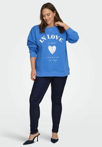 Sudadera azul de cuello redondo con un gráfico de corazón blanco y texto. Combinada con jeans ajustados oscuros y tacones azules de punta. Tejido suave, corte relajado.