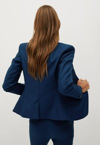 Blazer bleu marine ajusté avec épaules structurées, dos à basque et poignets à boutons. Le tissu est lisse avec une finition élégante.