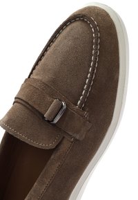 Mocassino in suede marrone con punta rotonda, caratterizzato da un design cucito, una fibbia in metallo argentato e una suola in gomma bianca.