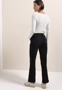 Femme aux cheveux courts marron porte un haut blanc à manches longues, un pantalon noir et des baskets blanches, debout les mains dans les poches, de profil.