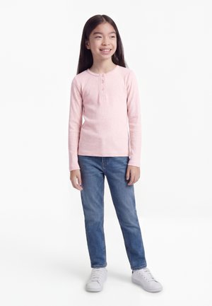 Chica sonriente con largo cabello oscuro, vestida con una blusa rosa de manga larga, jeans azules y zapatillas blancas sobre un fondo blanco.