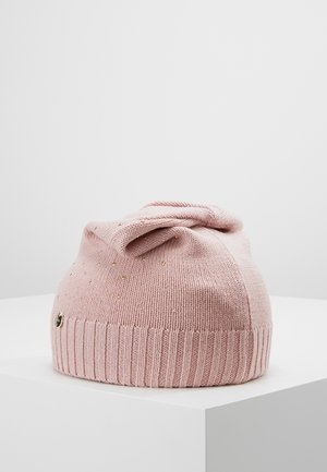 Gorro de punto color rosa claro con pequeños remaches dorados y un nudo decorativo en la parte superior, colocado sobre una superficie blanca.