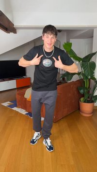 Νεαρός άνδρας με καθημερινά ρούχα που δείχνει το σχέδιο στη μαύρη μπλούζα του, όρθιος σε ένα σαλόνι με μεγάλο φυτό και πορτοκαλί καναπέ.