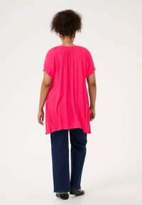 Tunica leggera di colore rosa acceso, con maniche corte, schiena plissettata e orlo più lungo, abbinata a pantaloni larghi blu scuro e ballerine nere.