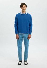 Jeune homme debout, portant un pull bleu sur une chemise rayée, un jean bleu clair et des mocassins noir et blanc, contre un fond clair uni.