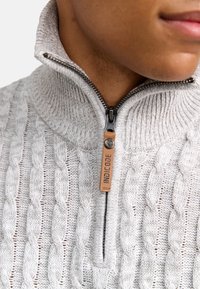 Grauer Strickpullover mit Reißverschlusskragen, der ein Zopfmuster aufweist und einen Lederaufnäher mit der Aufschrift "INDICODE" besitzt. Strukturiertes, mittelschweres Material.