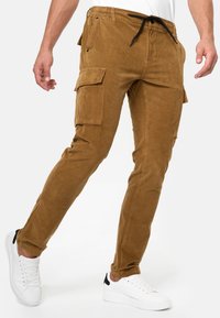 Braune Corduroy-Cargo-Hosen mit schmaler Passform, elastischem Bund, Kordelzug und seitlichen Taschen, kombiniert mit weißen Sneakern.