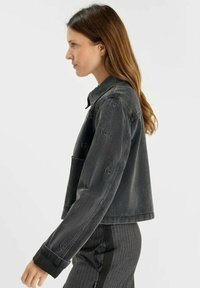 Chaqueta de mezclilla gris con un corte cropped, cuello y un sutil logotipo en las mangas. Textura suave con detalles de costura visibles.