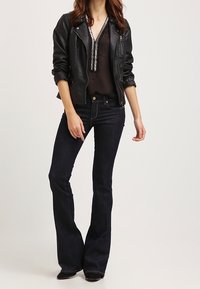 Donna che indossa una giacca di pelle nera, una camicetta nera trasparente con rifiniture argentate e jeans svasati scuri, in piedi davanti a uno sfondo bianco.