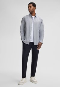 Camicia grigia con bottoni sopra una t-shirt bianca, abbinata a pantaloni scuri e sneakers bianche. Presenta una tasca sul petto e polsini con bottoni.