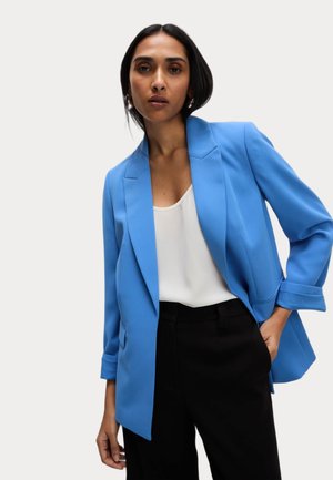 Blazer bleu clair au design ajusté, featuring un col en pointe, deux poches avant et des manches repliées, associé à un haut blanc et un pantalon noir.