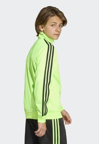 Neongrön träningsjacka med hög krage, svarta ränder längs ärmarna, mjukt material och ribbade muddar. Bärs tillsammans med matchande shorts.