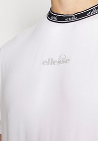 Camiseta blanca con cuello redondo, que presenta un logotipo en blanco y negro en el pecho y un cuello negro con acentos y marcas en blanco.