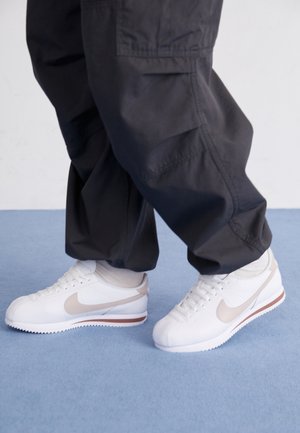 Personne portant des baskets Nike blanches et beiges avec un swoosh beige et un pantalon cargo noir sur un sol bleu clair.