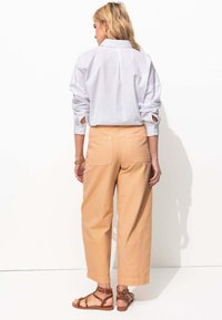Chemise légère blanche à manches retroussées, associée à un pantalon ample taille haute beige et à des sandales brunes à lanières.