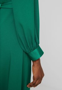 Une main à la peau foncée repose à côté d'une robe verte à manches longues avec un poignet à revers sur un fond neutre.