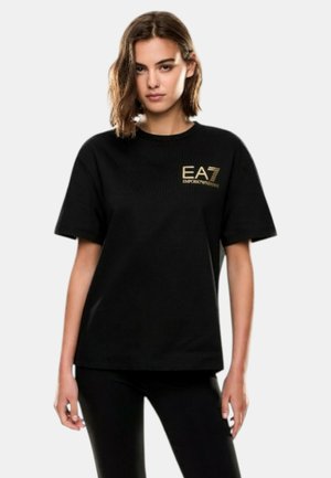 Giovane donna con capelli fino alle spalle, in piedi, che indossa una T-shirt nera EA7 Emporio Armani e pantaloni neri su uno sfondo chiaro e uniforme.