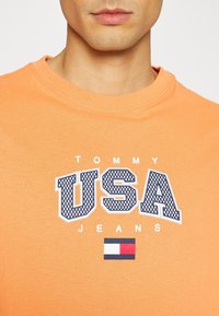 Oranžové bavlněné tričko s výrazným textem "USA" v námořnické modři a texturovaným vzorem, pod nímž se nachází malé logo v červených, bílých a modrých pruzích.