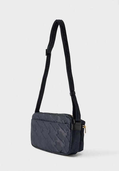 Μικρή ορθογώνια μαύρη τσάντα χιαστί με ρυθμιζόμενο λουρί, διακριτικό μοτίβο σε τονικότητα και χρυσή λεπτομέρεια φερμουάρ, τοποθετημένη σε απλό φόντο.