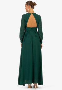Langes grünes Kleid mit transparenten langen Ärmeln, einem Schlüssellochausschnitt hinten und gerafftem Taillenbereich. Glatte Textur mit einem fließenden Rock.