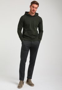 Sudadera verde oscuro con logo en relieve, bolsillo tipo canguro; combinada con pantalones de textura gris carbón y botas de tobillo marrón claro.