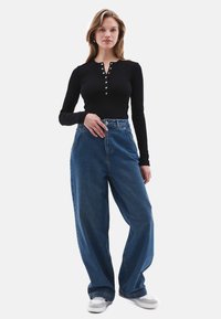 Schwarzes Langarmoberteil mit Knopfverschluss, kombiniert mit weiten blauen Denim-Jeans. Das Modell trägt weiße Sneakers. Schlichter Hintergrund.