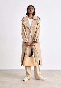 Beige trenchcoat van textuurstof, dubbelzijdige knopen, met biais in de zijkanten, gecombineerd met een crèmekleurige broek en een beige tas. Witte top en sneakers.
