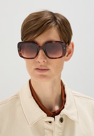Persona con capelli corti castani che indossa grandi occhiali da sole quadrati in tartaruga e una giacca beige sopra una camicia marrone.