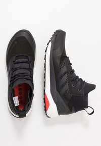 Chaussures de sport noires avec une tige en mesh, des détails texturés et un marquage trois bandes bien visible. Semelle intercalaire blanche avec des accents rouges.