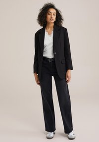 Fekete oversized blézer egy csipkés szélű fehér felsőn, magas derékú fekete farmernadrággal és fekete-fehér tornacipővel párosítva.