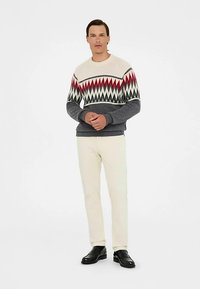 Gestrickter Pullover mit cremefarbenem Grundton und rotem sowie schwarzem Zickzackmuster, graue Ärmel, ergänzt durch cremefarbene Hose und schwarze Schuhe.