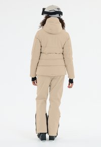 Beige pufferjacka med huva, utsvängda matchande byxor och svarta skidboots. Anmärkningsvärda räfflade detaljer och blixtlås i byxans nederkant.