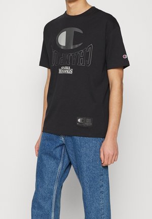 Sort bomulds-T-shirt med stort hvidt tryk med teksten "Champion" og "Stranger Things." Indeholder et logo-patch nær kanten.