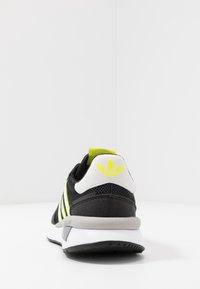 Chaussure de sport noire avec des bandes jaunes, empeigne en mesh, talon blanc et semelle en caoutchouc, avec un logo à l'arrière. Design simple et fonctionnel.