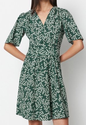 Woman wearing a green dress with white abstract dot pattern, short sleeves, V-neckline, and a fitted waist standing against a plain background.