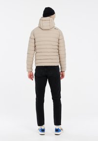 Doudoune beige avec un matelassage horizontal, capuche attachée et logo dans le haut du dos, associée à un jean noir et des baskets multicolores.