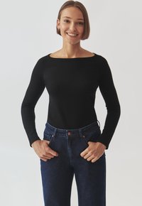Top lung cu mâneci lungi, de culoare neagră, cu guler tip barcă, asortat cu blugi din denim albastru închis, cu talie înaltă. Materialul pare moale și aderent.