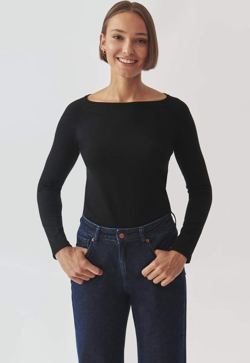 Top lung cu mâneci lungi, de culoare neagră, cu guler tip barcă, asortat cu blugi din denim albastru închis, cu talie înaltă. Materialul pare moale și aderent.