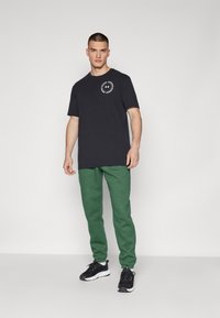 Sort bomulds T-shirt med et logo på brystet, parret med grønne fleece sweatpants. Modellen har sorte træningssko på. Neutral baggrund.