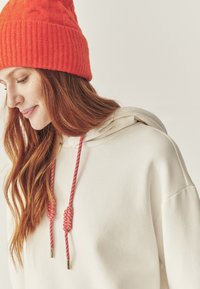 Sudadera blanca con cordón que presenta acentos trenzados en rosa y púrpura con puntas metálicas. Un gorro de punto naranja complementa el atuendo.