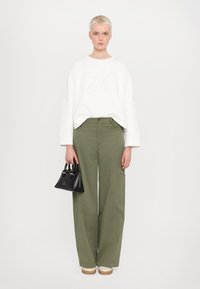 Sweat-shirt blanc avec lettrage en relief, pantalons larges vert olive, sac à main noir et chaussures à plateforme crème sur fond uni.
