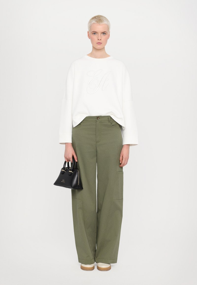 Sweat-shirt blanc avec lettrage en relief, pantalons larges vert olive, sac à main noir et chaussures à plateforme crème sur fond uni.