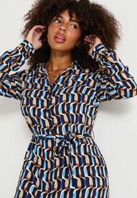 Vestido abotonado con un patrón geométrico en azul, marrón y beige. Mangas largas con puños de botón y detalle de cintura atada. Textura de tela suave.