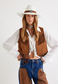 Gilet in suede marrone con frange sopra una blusa bianca, abbinato a jeans in denim blu con accenti in suede marrone, borchie e un cappello bianco con una fascia marrone.