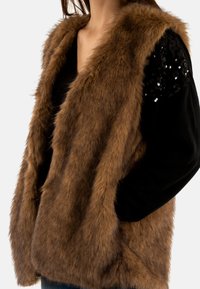 Gilet in finta pelliccia marrone con fit rilassato, apertura frontale e maniche accentuate da paillettes nere. Texture morbida e design leggero.