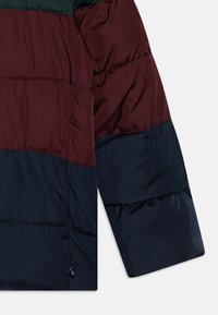 Μπουφάν με επένδυση που διαθέτει σχέδιο χρωματικών αποκλεισμών σε μπορντό, πράσινο και ναυτικό μπλε. Λείες υφές με εμφανή ραφή και λογότυπο στην προεξοχή.