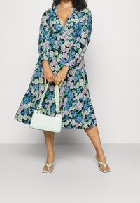 Robe fleurie avec un décolleté en V profond, des manches longues et un ourlet en couches. Présente des fleurs bleues et violettes sur un fond noir. Associée à un sac vert menthe.
