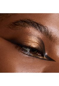 Gros plan sur un œil présentant un eyeliner doré métallisé et noir intense. La peau a une teinte chaude et lisse, avec des cils et des sourcils bien définis.