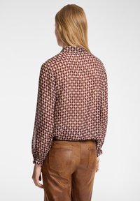 Blusa trasparente color crema con motivo geometrico bordeaux, maniche lunghe con polsini arricciati e colletto. Indossata con pantaloni in pelle marrone.